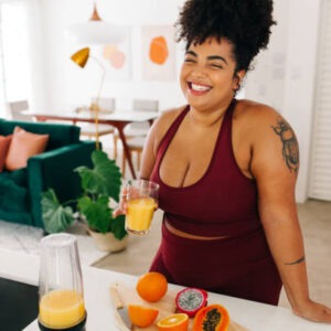 happy woman holding a glass of fresh juice in the kitchen while chopping up fruits for her weight loss goals