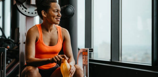 Beautiful fitness woman dressed up in sportswear sitting at gym resting after workout and thoughtfully looking into the distance holding a water bottle